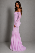 A woman modeling a lavender off-the-shoulder maxi dress with long lace sleeves and a flared mermaid skirt, photographed against a plain gray studio backdrop.