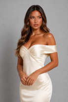 A woman modeling an off-the-shoulder champagne satin dress against a neutral gray studio background.