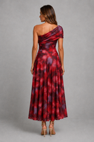 A woman shown from the back wearing a one-shoulder maxi dress with a red, burgundy, and purple tie-dye pattern, paired with beige high-heeled sandals, against a neutral gray studio background.
