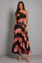 A woman modeling a one-shoulder maxi dress with a black and dusty rose tie-dye pattern, standing against a neutral gray studio background.