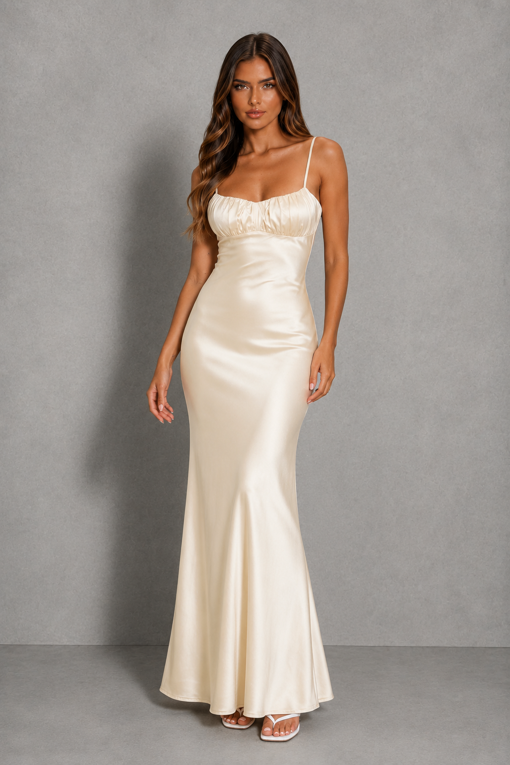 A woman modeling a floor-length ivory satin slip dress with spaghetti straps and a gathered sweetheart neckline, standing against a neutral gray studio backdrop.