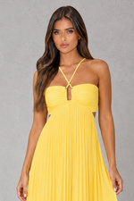 A woman modeling a bright yellow strapless maxi dress with a sweetheart neckline, thin crisscross halter straps, and a pleated skirt, set against a neutral gray studio background.