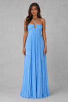 A woman modeling a light blue strapless maxi dress with a pleated skirt and a keyhole cutout at the bust, standing against a neutral gray studio background.