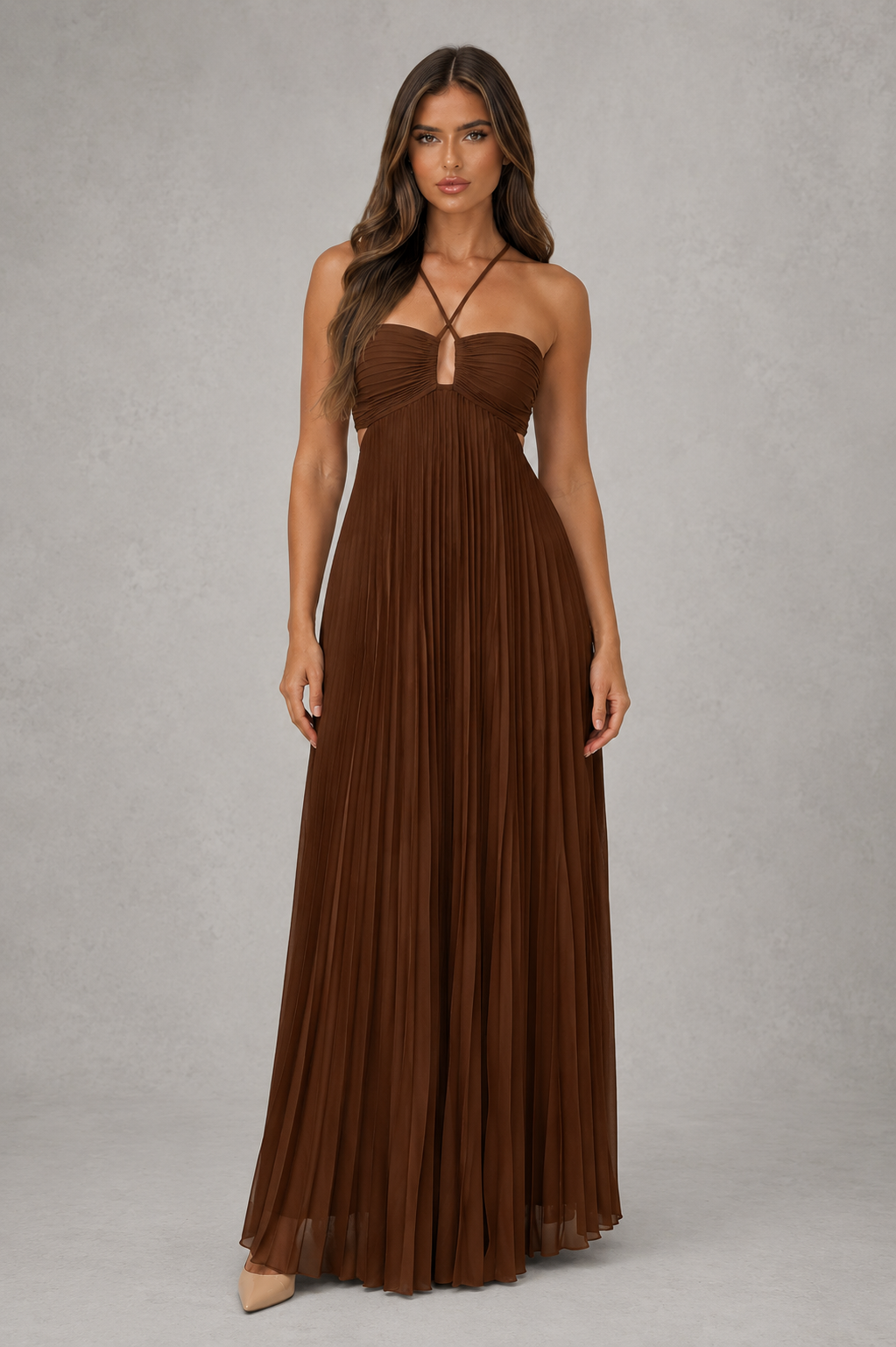 A woman modeling a floor-length brown pleated maxi dress with a strapless sweetheart neckline and thin crisscross tie details at the chest, photographed against a neutral gray studio backdrop.