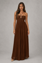 A woman modeling a floor-length brown pleated maxi dress with a strapless sweetheart neckline and thin crisscross tie details at the chest, photographed against a neutral gray studio backdrop.