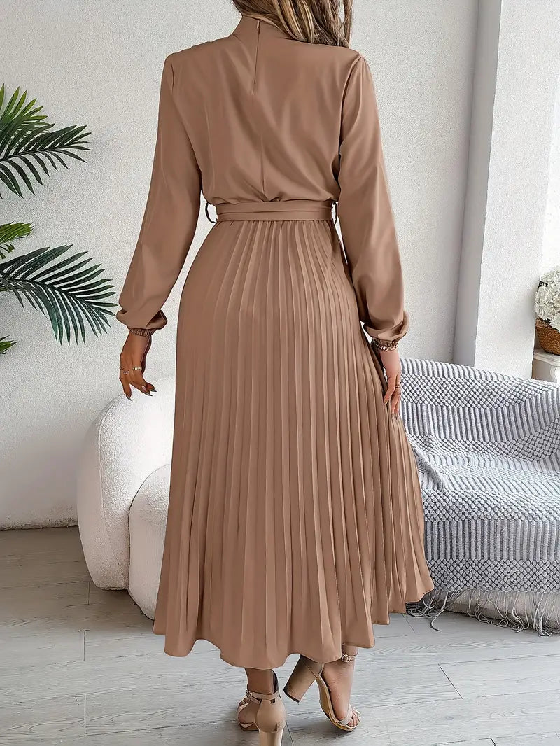 Woman wearing a brown pleated dress in a room with a white wall and a plant.