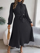 Woman wearing a black dress with long sleeves in a room with a white wall and a plant.