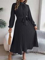 Woman wearing a black dress with long sleeves in a room with a white wall and a plant.
