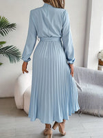 Woman wearing a light blue pleated dress in a room with a couch and plant.