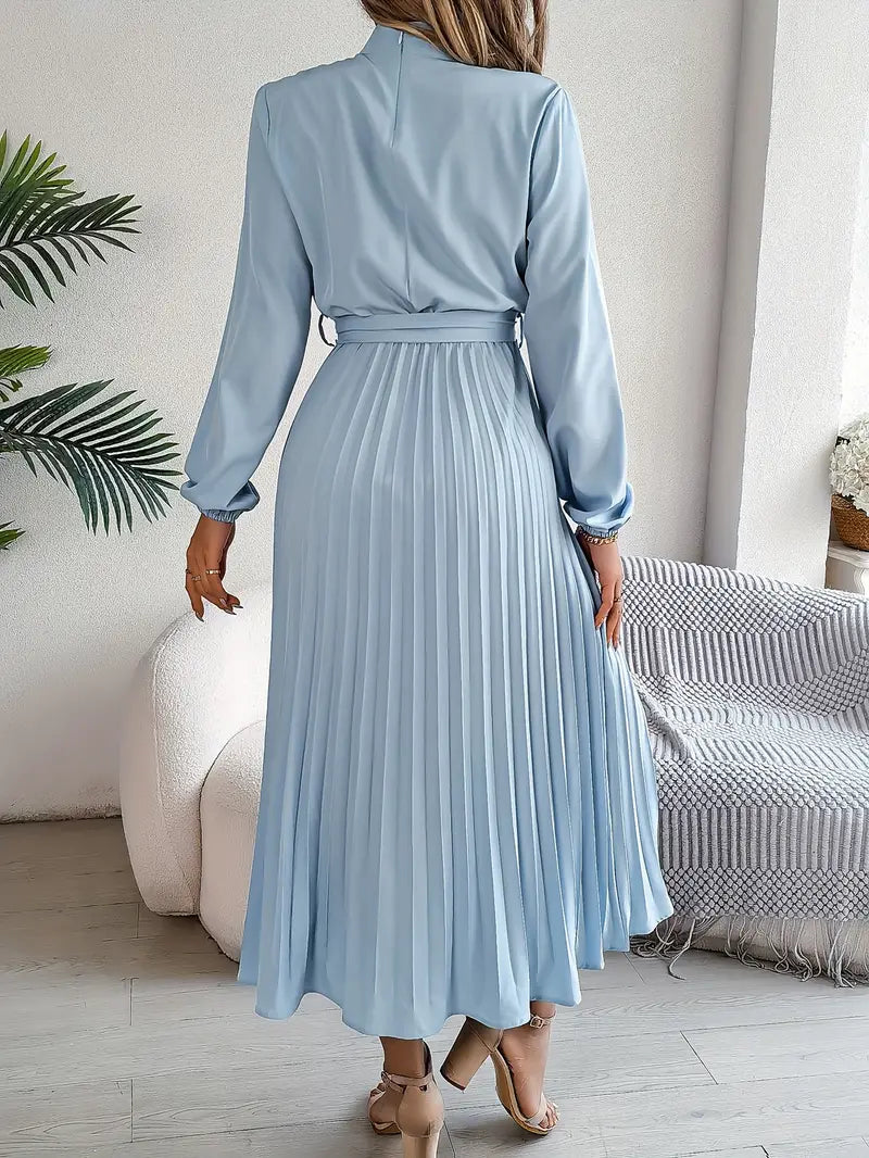 Woman wearing a light blue pleated dress in a room with a couch and plant.