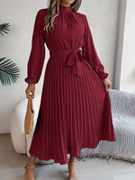 Woman wearing a burgundy pleated dress in a room with a couch and plant.