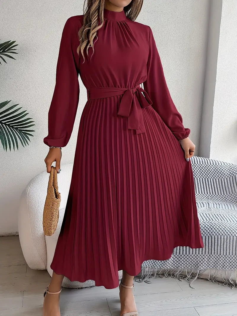 Woman wearing a burgundy pleated dress in a room with a couch and plant.