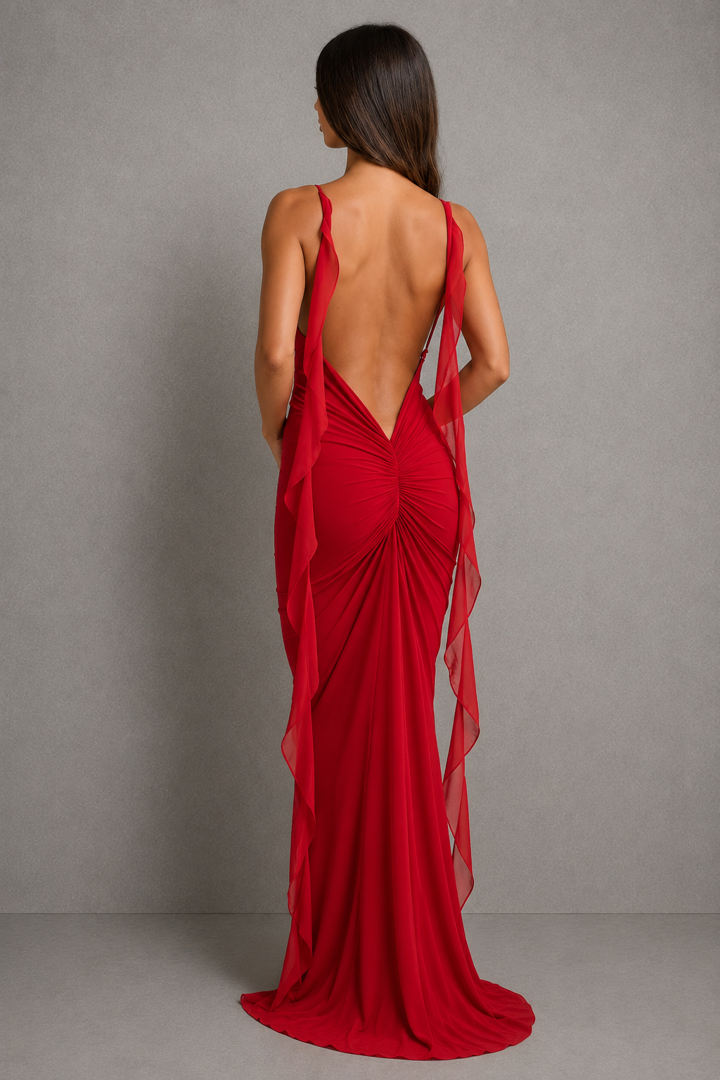 A woman wearing a long red evening gown shown from the back against a plain gray studio backdrop. The dress has a deep open back, thin straps, gathered draping fabric, and a flowing train.