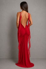 A woman wearing a long red evening gown shown from the back against a plain gray studio backdrop. The dress has a deep open back, thin straps, gathered draping fabric, and a flowing train.