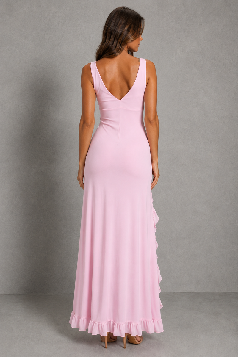 A woman modeling a light pink sleeveless maxi dress from the back, featuring a deep V-neckline, fitted bodice, and cascading ruffles along the side and hem, set against a neutral gray studio backdrop.