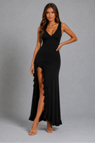 A woman modeling a black sleeveless maxi dress with a deep V-neckline, fitted silhouette, and a high ruffled thigh slit, photographed against a plain gray studio background.