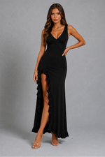 A woman modeling a black sleeveless maxi dress with a deep V-neckline, fitted silhouette, and a high ruffled thigh slit, photographed against a plain gray studio background.
