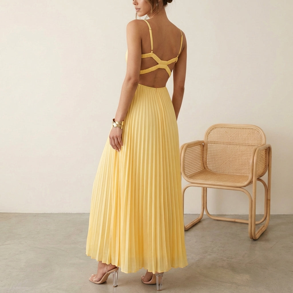 Woman wearing a yellow pleated dress standing next to a wicker chair against a plain background