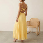 Woman wearing a yellow pleated dress standing next to a wicker chair against a plain background