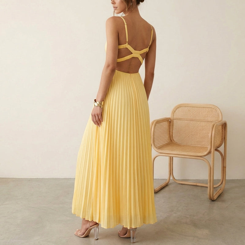 Woman wearing a yellow pleated dress standing next to a wicker chair against a plain background