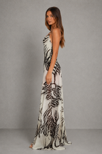 A woman modeling a sleeveless, floor-length maxi dress with a black and white zebra-like animal print, standing against a textured gray studio backdrop.