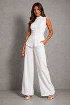 A woman modeling a white sleeveless peplum top and matching wide-leg trousers set, standing against a textured gray studio background.