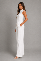 A woman modeling a white sleeveless high-neck jumpsuit with wide-leg pants and side pockets, standing against a neutral gray studio backdrop.