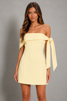 A woman modeling a pale yellow off-the-shoulder mini dress with a wide draped band across the chest and a long tie detail on one side, photographed against a plain gray studio background.