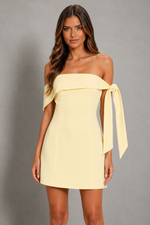 A woman modeling a pale yellow off-the-shoulder mini dress with a wide draped band across the chest and a long tie detail on one side, photographed against a plain gray studio background.