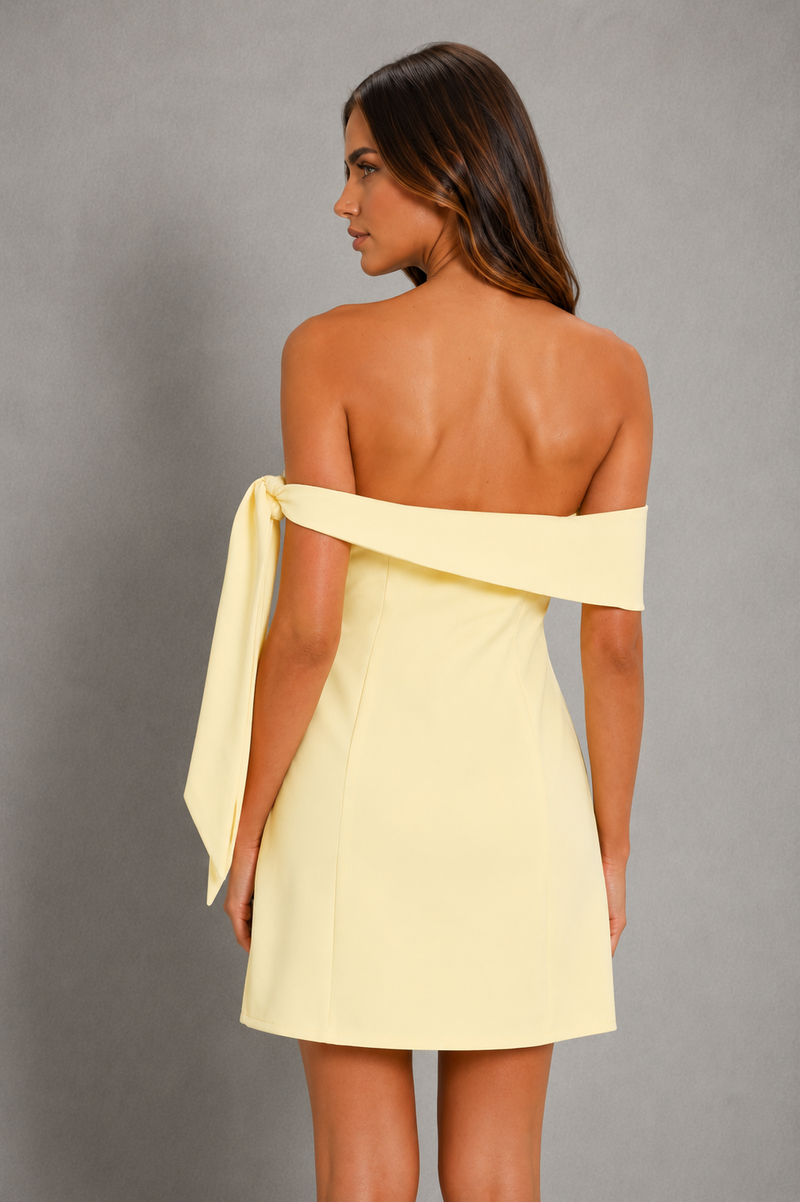 A woman shown from the back wearing a pale yellow off-the-shoulder mini dress with a wide draped strap, photographed against a plain gray studio background.