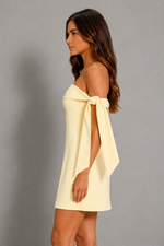 A woman wearing a pale yellow off-the-shoulder mini dress with a large bow detail at the neckline, shown in profile against a plain gray studio background.