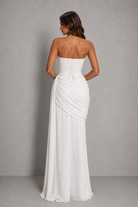 A woman modeling a strapless white floor-length gown from the back, showing a lace-up corset-style back and draped skirt, against a plain gray studio background.