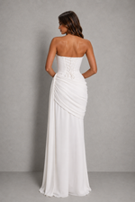A woman modeling a strapless white floor-length gown from the back, showing a lace-up corset-style back and draped skirt, against a plain gray studio background.