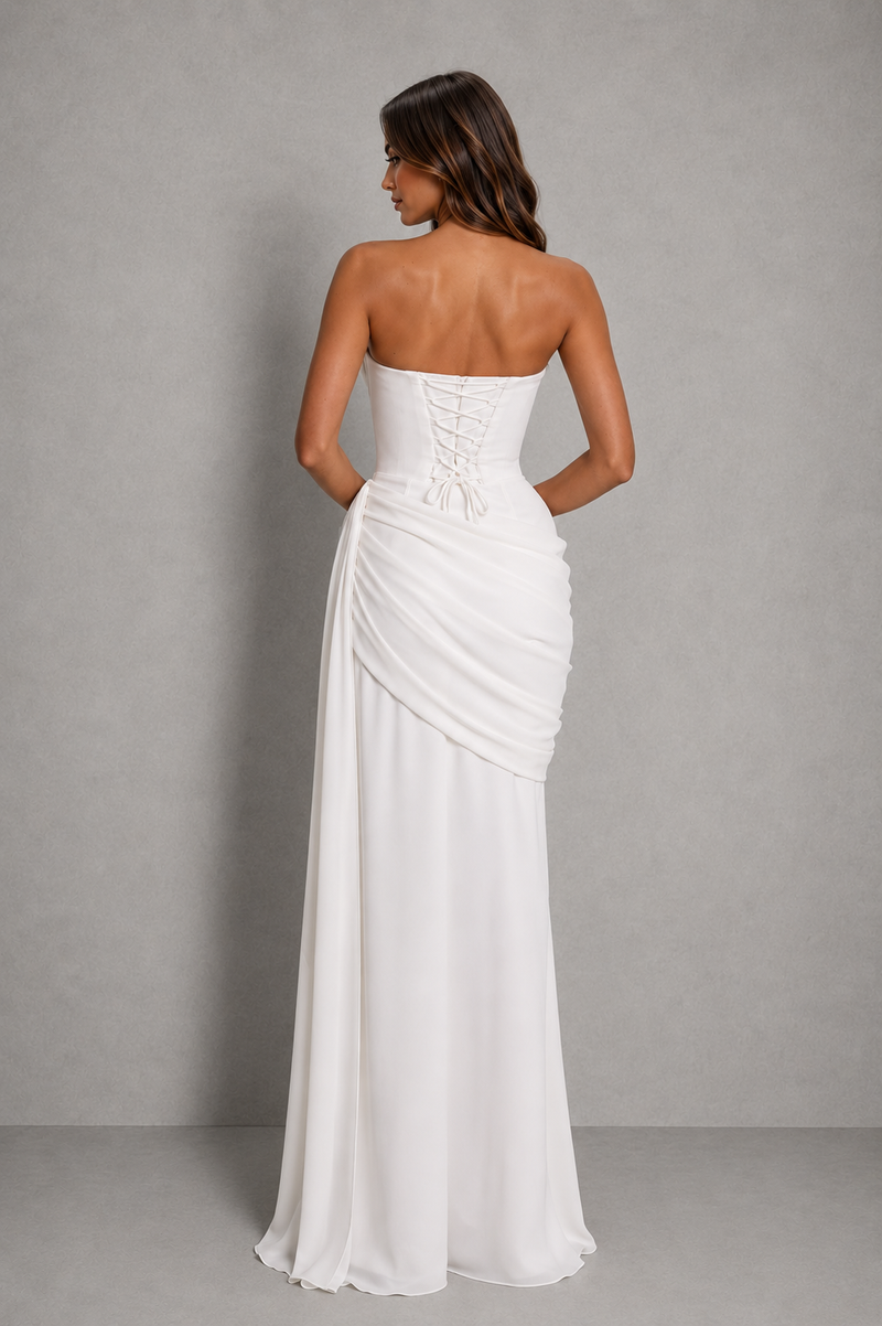 A woman modeling a strapless white floor-length gown from the back, showing a lace-up corset-style back and draped skirt, against a plain gray studio background.