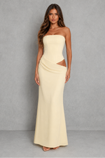 A woman modeling a strapless, floor-length cream maxi dress with a structured bodice, side cutouts, and a draped skirt, set against a textured gray studio backdrop.