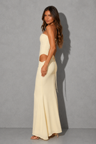 A woman modeling a cream-colored two-piece set consisting of a strapless crop top and a matching high-waisted maxi skirt with side cutouts and tie details, set against a gray studio background.