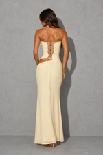 A woman stands with her back to the camera wearing a strapless cream maxi dress with a lace-up corset-style back and a side cutout, photographed against a gray studio backdrop.