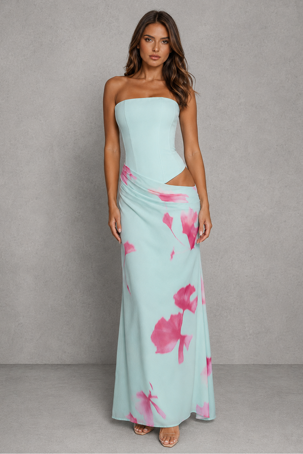 A woman modeling a strapless light blue maxi dress with pink floral watercolor print and side cutouts, standing against a textured gray studio backdrop.