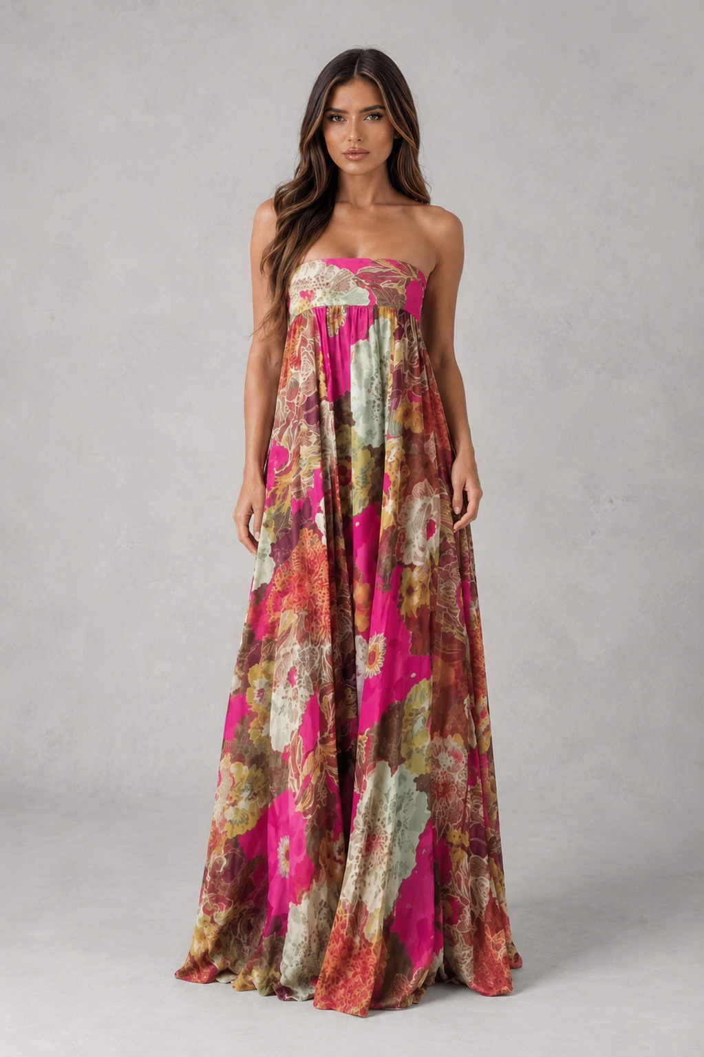 A woman modeling a strapless maxi dress with a vibrant abstract floral print in pink, orange, and beige tones against a neutral gray studio background.