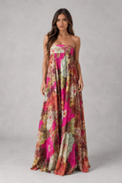 A woman modeling a strapless maxi dress with a vibrant abstract floral print in pink, orange, and beige tones against a neutral gray studio background.