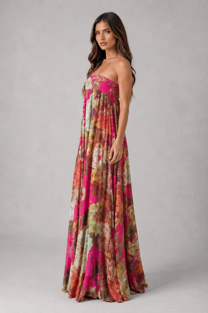 A woman modeling a strapless maxi dress with a vibrant abstract floral print in pink, orange, and beige tones, set against a neutral gray studio background.