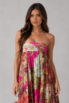 A woman modeling a strapless maxi dress with a vibrant floral print in pink, green, and brown tones against a neutral gray studio background.