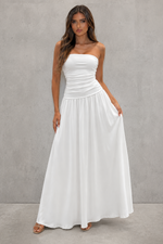 A woman modeling a strapless white maxi dress with a ruched bodice and flowing skirt, standing against a textured gray studio wall.