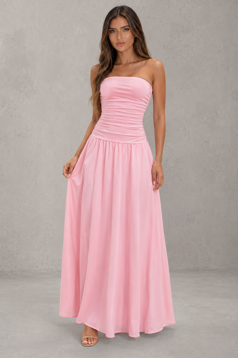 A woman modeling a strapless pink maxi dress with a ruched bodice and flowing skirt, standing against a textured gray studio backdrop.