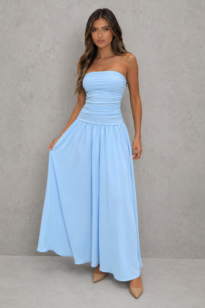 A woman modeling a strapless light blue maxi dress with a ruched bodice and flowing skirt, standing against a textured gray studio wall.