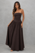 A woman modeling a strapless, floor-length dark brown maxi dress against a textured gray studio backdrop.
