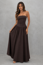 A woman modeling a strapless, floor-length dark brown maxi dress against a textured gray studio backdrop.
