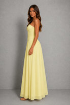 A woman modeling a pale yellow strapless maxi dress with a ruched bodice and flowing skirt, standing against a neutral gray studio backdrop.