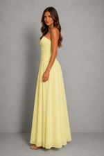 A woman modeling a pale yellow strapless maxi dress with a ruched bodice and flowing skirt, standing against a neutral gray studio backdrop.
