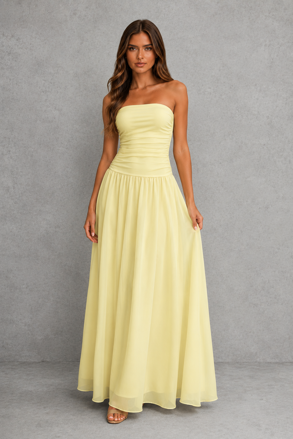 A woman modeling a strapless pale yellow maxi dress with a ruched bodice and flowing skirt, standing against a textured gray studio backdrop.